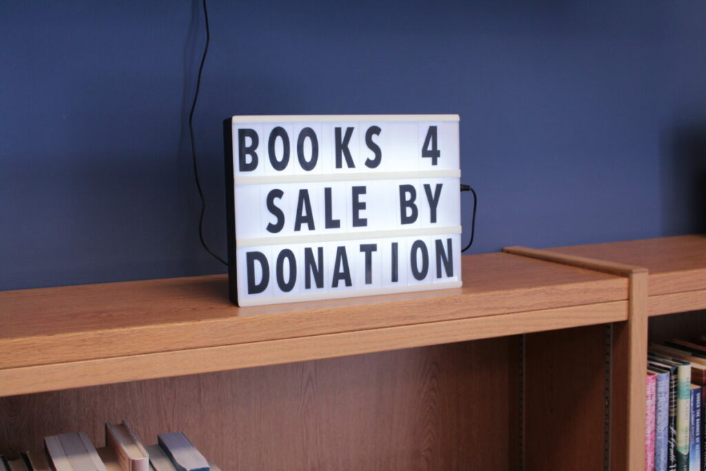 A sign on a bookshelf reads "BOOKS 4 SALE BY DONATION"