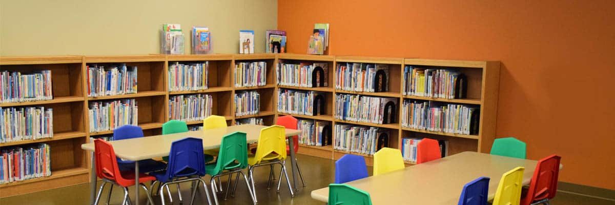 A library early reader area with two tables surrounded by rainbow chairs and bookshelves with early reader books.
