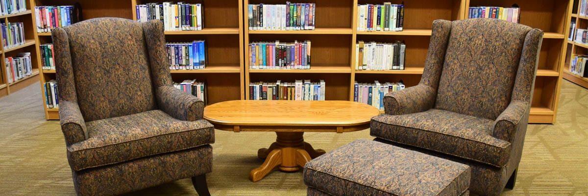 A library reading area with two armchairs, an ottoman and bookshelves in the background.