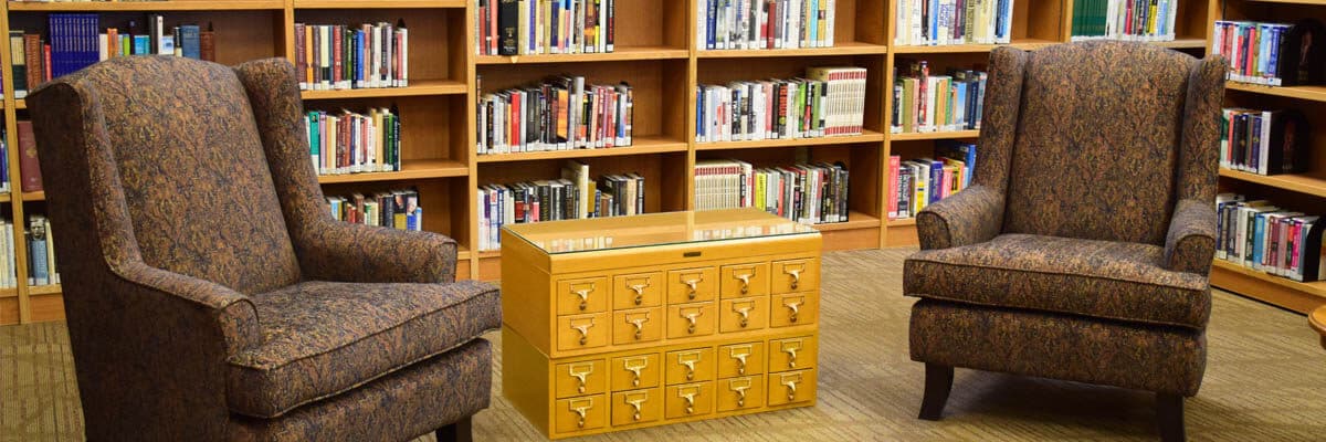 A reading area in a library with two armchairs, a card catalog coffee table and bookshelves in the background.