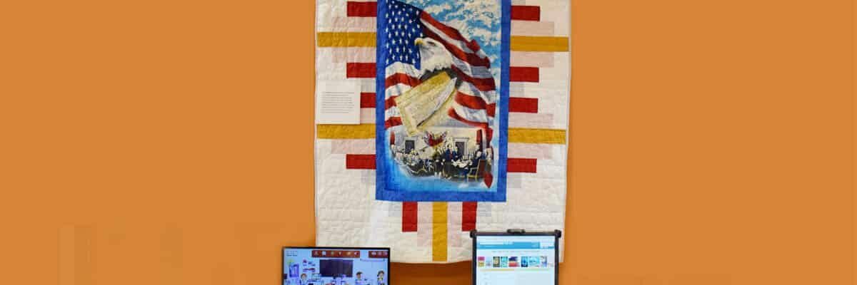 Quilt with an American flag and eagle hanging above two computer monitors on an orange wall.